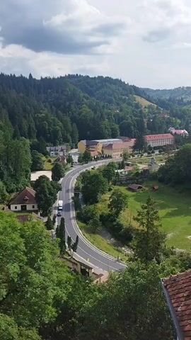The video pans across a scenic landscape. In the foreground, the terracotta tiled roof of a building is visible, with a white chimney. Beyond the roof, a winding road curves through a lush green valley dotted with trees. On either side of the road, houses and larger buildings, including a long, multi-story structure with a red roof, are nestled amongst the trees. The valley is surrounded by rolling hills covered in dense green forest, with patches of grass and smaller buildings visible on the slopes. The sky is partly cloudy. The overall impression is of a rural or semi-rural area with a mix of natural beauty and human habitation.