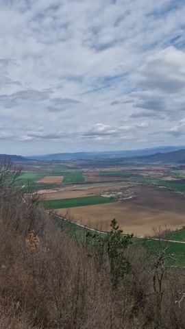 The video shows a panoramic view of a valley from a high vantage point. The foreground is framed by bare trees with some green vegetation emerging, suggesting early spring. The landscape stretches out to a broad valley composed of agricultural fields in various shades of green and brown, indicating different crops or stages of cultivation. A road meanders through the valley.  In the distance, a mountain range is visible, with some peaks capped with snow. The sky is partially cloudy, with patches of blue sky peeking through. A blue railing is visible at the edge of the frame in the lower right corner, suggesting that the viewpoint is a man-made structure. There are no people visible in the video.