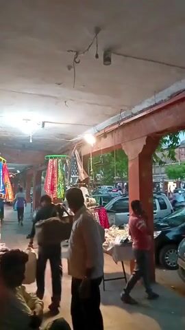 The video shows a market area during the day. Several people are present, including shoppers and vendors. One man in a pink shirt talks on his cell phone. A man in a white shirt carries a large bag. There are multiple cars parked nearby, and traffic is visible. A building with arched openings can be seen in the background. The market area is covered by a ceiling with exposed electrical wires and light fixtures.