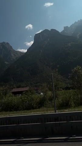 The video shows a view from a moving vehicle. The scene is dominated by a large, dark mountain range under a clear blue sky with a few scattered clouds. The lower slopes of the mountains are covered in dense green trees. In the foreground, there is a road with a guardrail, and more trees and vegetation. A tall, thin pole is visible on the right side of the frame. Power lines are stretched across the sky. There are no people or specific actions visible. The overall impression is a scenic view of a mountainous landscape during the daytime.