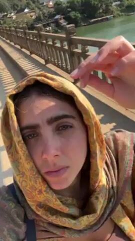 A woman wearing a yellow paisley headscarf stands on a bridge with ornate concrete railings. She looks directly at the camera, puckering her lips slightly. Behind her, a wide river reflects the bright sky, and a green hillside rises in the background. Houses are visible on the riverbank. The day is sunny and bright.
