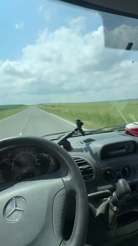 The video captures a first-person perspective from the driver's seat of a Mercedes-Benz van or truck, driving along a straight, two-lane asphalt road. The driver's right hand is visible resting on the steering wheel, which features the prominent Mercedes-Benz logo.

The dashboard is visible, showing the speedometer and tachometer, with the vehicle's speed appearing to be between 80 and 90 km/h. A phone holder is mounted on the dashboard, and the gear stick is visible to the right.

Outside the vehicle, the road extends far into the distance, flanked by extensive green fields and some trees or bushes on the horizon. The sky is bright blue with scattered white, fluffy clouds, indicating a sunny day. Through the side mirror, the passing green landscape and part of the white side of the vehicle are visible.

The scene depicts a journey through open, rural countryside, consistent with the location context of Madara, Bulgaria. The audio includes the sound of the vehicle moving, suggesting constant forward motion.