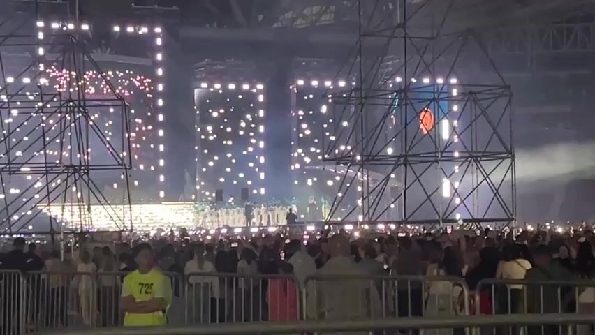 The video shows a large outdoor concert in a stadium. The stage is set up with multiple screens displaying bright lights, and there are large metal scaffolding structures on either side. A crowd of people is gathered in front of the stage, many holding up their phones with lights on. Some security personnel can be seen in the foreground. The stadium seating is visible in the background, with some sections appearing to be red and white. Smoke or fog is present on the stage, illuminated by the lights. The overall atmosphere appears to be energetic and festive.