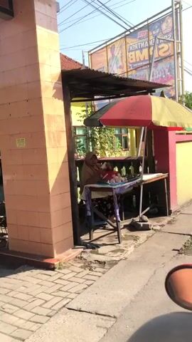 A woman wearing a hijab and a brown hijab is sitting at a small stall selling food. She is holding a piece of food and appears to be eating it. There are several plates of food on the stall. The stall is located outdoors, next to a building with yellow walls and a red roof. There is a sidewalk in front of the stall, and a street to the right. A large billboard is visible in the background. The sky is blue and there are some clouds.