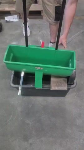 A person wearing khaki shorts and white sneakers is standing behind a green broadcast spreader. The spreader has a black plastic tray beneath it and is resting on a wooden block. The person is holding the spreader's handles, and their right hand is adjusting the spreader. The background shows a concrete floor and some wooden pallets. The lighting is overhead and appears to be artificial.