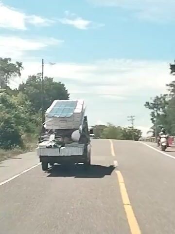The video shows a rear view of a flatbed truck loaded with cargo driving on a paved road. The truck is on the right side of the road, and the camera is positioned behind and slightly to the left of the truck, also on the road. The truck's bed is piled high with boxes, some stacked vertically and others horizontally. The boxes appear to be light-colored, possibly white or cream, with some text or logos visible. On top of the boxes, there are also some blue, possibly transparent, sheets, which could be plastic sheeting or perhaps solar panels. Bundles of what look like pipes or metal rods are also visible, secured to the truck bed. A white hard hat is perched on top of some of the cargo. The truck is moving forward, and its shadow is cast on the road behind it. The road itself is asphalt with a yellow line marking the center. On the left side of the road, there are trees and foliage. On the right side, beyond a concrete barrier, there are more trees and utility poles. The sky is blue with scattered white clouds, suggesting a daytime scene with clear weather. The overall impression is that of a commercial or delivery vehicle transporting goods.