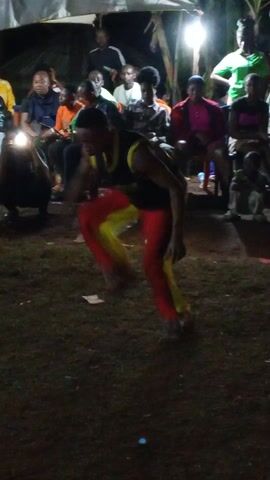 In a dimly lit outdoor setting, a young man dressed in a black tank top, red pants with yellow panels, is performing an energetic dance in front of a seated audience. The audience, comprising mainly young adults, are watching the performance attentively, some with phone flashlights directed at the dancer. They are seated on chairs, arranged in rows under what appears to be a makeshift tarp shelter. The ground is covered in grass and some debris. The scene suggests a casual, possibly informal, cultural or entertainment event taking place at night.