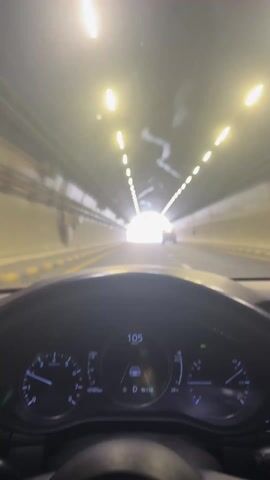 The video shows a car driving through a tunnel and then emerging into bright daylight. 

The viewpoint is from the driver's seat. The car is moving forward at around 104 kilometers per hour. The dashboard is visible, showing the car's speed and other information. The tunnel has bright lights overhead, arranged in rows. Another car is visible ahead.

As the car exits the tunnel, the view opens up to a clear blue sky and a landscape of brown mountains with white cliffs. The road is a multi-lane highway with yellow lines separating the lanes. There are road signs indicating a speed limit of 80. Other cars are visible in the distance.