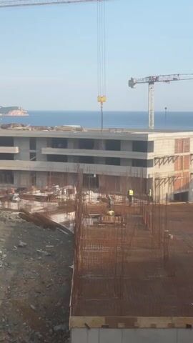This media file captures a construction site under a clear blue sky, suggesting a pleasant day. The primary subjects are several construction workers, distinguished by their bright yellow safety vests, diligently working on a multi-story building. They are engaged in construction activities, likely involving the placement of reinforcement bars and concrete pouring, as evidenced by the extensive network of rebar visible throughout the structure.

The scene is dominated by a large, modern building in various stages of construction. The lower floors appear to be more complete, with concrete structures and brickwork visible, while the upper levels are still primarily reinforced concrete frameworks with visible rebar. Two prominent cranes are present: a taller, lighter-colored tower crane in the background and a smaller, yellow hook assembly hanging from cables in the foreground, indicative of ongoing lifting operations.

In the distance, a body of water, likely the Adriatic Sea given the location context of Boreti, Montenegro, is visible. A coastal landmass or island can be seen on the horizon, adding to the scenic backdrop. The time of day appears to be morning or late afternoon, judging by the angle of the sun and the shadows cast. The overall mood is one of industrial activity and progress, with the construction work juxtaposed against the natural beauty of the coastal environment. No visible text is present in the media file.