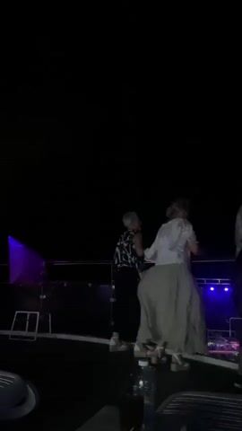 The video shows a nighttime scene of people dancing on what appears to be a boat or deck. The lighting is dim with occasional flashes of colored lights, primarily purple and blue, illuminating the dancers. Two women are visible dancing. One woman, on the left, wears a black and white patterned top and dark pants. The other woman, in the center, wears a white top and a long, flowing light-colored skirt. Both are facing away from the camera, engaged in movement. A pink flag is visible on a pole to the left of the frame. In the foreground, out of focus, are parts of chairs and a table with a bottle and a glass. The background is dark, suggesting it is night and there is no land visible. The overall atmosphere appears to be a lively outdoor gathering or party.