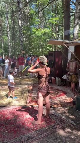 A person in a brown striped outfit with a floral headpiece is hooping with a green hula hoop in front of a group of people in a wooded area. The person is standing on a red patterned rug. To the left of the person, a young boy in a brown outfit is watching. Behind the person is a white trailer with speakers, and several adults are standing around. The ground is covered in leaves and dirt. The scene appears to be outdoors during the day.