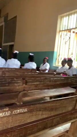 The video shows a group of people, likely students or trainees, sitting in a room with wooden pews. They are mostly wearing white uniforms with red accents on their caps, suggesting a medical or nursing profession. The room appears to be a hall or chapel, with a window letting in natural light and a high ceiling with fans and light fixtures. Some individuals are facing forward, seemingly listening or observing something, while others have their heads bowed. The pews are old and worn, with some inscriptions visible on them. The overall atmosphere seems solemn or attentive.