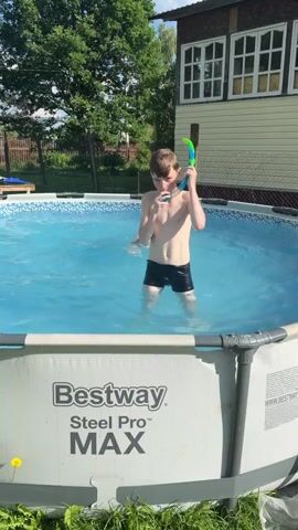 A young boy wearing swim trunks and snorkel gear stands in a Bestway Steel Pro MAX above-ground swimming pool. He dips his head underwater, then emerges and adjusts his snorkel. Another person, partially submerged, is visible in the background. The pool is set up in a yard with green grass and trees surrounding it, and a building with a light-colored exterior and windows is visible behind the pool. The sky is bright and the lighting suggests it is daytime.
