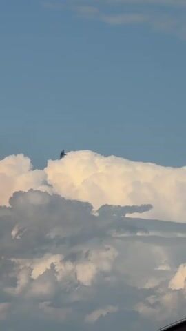 The video shows a blue sky with white clouds. A bird is flying in the distance above a large, puffy cloud. The bottom of the frame shows the tops of buildings.