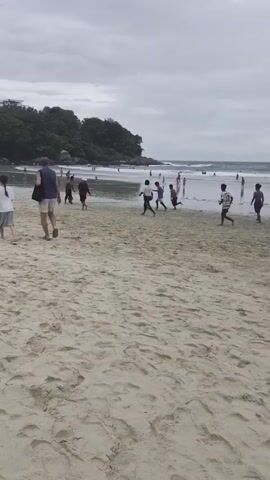 The video shows a sandy beach with people walking and playing in the distance. The sky is overcast, and there are trees and buildings on a hill in the background. The camera is focused on the sand in the foreground, with the beach and ocean further back. Several people are scattered across the beach, with some walking and others appearing to be playing a game. The overall mood is relaxed and casual.