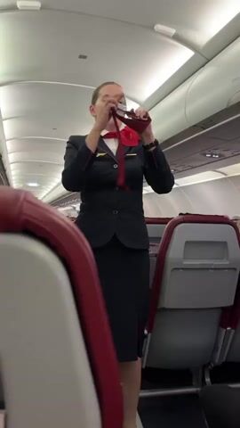 A flight attendant in uniform, consisting of a dark suit jacket, a white shirt, and a red scarf, stands in the aisle of an airplane. She is holding a red cloth, possibly a life vest or safety demonstration item, and appears to be demonstrating its use. The seats in the foreground and background are upholstered in red and grey. The overhead compartments and lighting fixtures of the airplane cabin are visible.