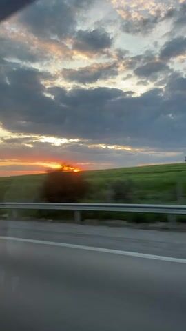 The video shows a sunset view through a car window. The sun is setting behind a green field, casting an orange glow on the sky. Some clouds are in the sky, some colored in hues of orange and pink from the sunset. A road and a metal guardrail are visible in the foreground.  A sign with an arrow points to "XO Park 16 km." The camera appears to be moving, as the scenery is changing. There is a power line in the distance.