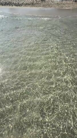 The video shows a first-person perspective of someone walking in shallow, clear water at a beach. The sun is shining brightly, creating dappled light patterns on the sandy bottom, indicating it's likely daytime and sunny weather. The water is very calm and transparent, allowing a clear view of the sand and any small debris or plants on the seabed.

The subject's feet are visible, one at a time, as they move through the water. The person is wearing sandals or flip-flops, and a patterned beach wrap or skirt is visible around their waist. The scene evokes a sense of relaxation and enjoyment associated with a beach vacation. The gentle movement of the water and the clear visibility suggest a peaceful and pleasant environment. No text is visible in the video. The location context provided is Santa Eulària des Riu, Spain, which is known for its beautiful beaches and clear waters, consistent with the visual cues in the video.