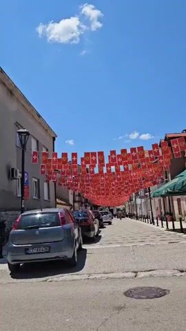 This video appears to be a street-level view of a town square or main street in Cetinje, Montenegro, likely during the daytime on a sunny day. 

The scene is characterized by buildings with a mix of architectural styles, some appearing older with traditional European features like balconies and awnings, while others are more modern. The street is paved, and there are parked cars visible, including a silver sedan, a red SUV, and a motorcycle. 

A prominent feature is a string of numerous small red flags with white crosses, hung across the street, suggesting a possible celebration, festival, or national event. These flags create a festive atmosphere.

There are no people clearly visible in the footage, nor is any specific activity or event actively taking place beyond the general ambiance of a town street.

Notable details include:
- A parking sign indicating "Zona 1" with additional text in what appears to be Montenegrin.
- The text "OSVIT" visible on a white awning of a shop.
- The text "STARA GARDA" on a sign for a business.
- The clear blue sky with a few white clouds, indicating good weather.
- The overall impression is one of a quiet, sunny day in a European town adorned for a special occasion.