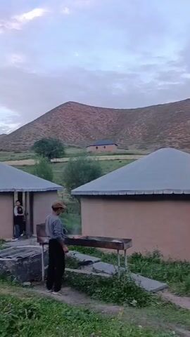 The video shows an outdoor scene with traditional dwellings, possibly yurts or similar structures, in a mountainous area. Two such circular structures with conical roofs are visible in the foreground. One is a light-colored yurt with some decorative elements and an open entrance. The other is a more robust structure with a beige wall and a darker grey roof. A person stands near the entrance of the beige structure, and another person is visible inside. In front of the beige structure, there is a long grill with what appears to be meat cooking on it, and a person in a baseball cap is standing beside it. In the background, hills or mountains rise, with some scattered trees and vegetation. The sky is cloudy and appears to be either early morning or late evening. The overall impression is that of a rural or encampment setting, possibly a tourist or nomadic site.