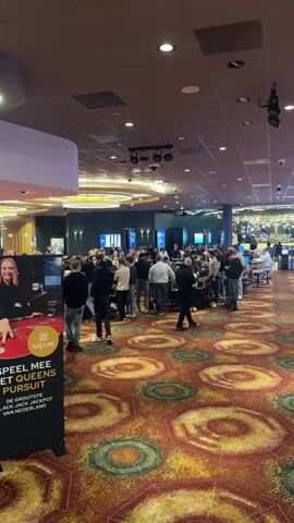 The video shows the interior of a casino.  The first shot has a group of people standing in a crowd around what appear to be gaming tables.  There is a sign visible on the left that is advertising "PEEL MEE MET QUEENS PURSUIT DE GROOTSTE BLACK JACK JACKPOT VAN NEDERLAND".  The camera then pans to the right showing rows of slot machines with some people playing them. There is a sign hanging on a column indicating "TICKETS & CASH". The carpet has a large round pattern with a few different colors that include orange, green and black. The ceiling is low with recessed lighting and surveillance cameras. There is also track lighting over the gaming tables.