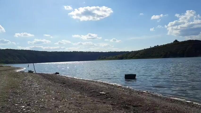 This media file depicts a scenic waterfront view at Sokil, Ukraine. The scene is dominated by a large body of tranquil blue water, reflecting the bright daytime sky dotted with fluffy white clouds. The sun is shining, suggesting a clear and pleasant day.

On the left side of the frame, a gravelly shore leads to the water's edge. Scattered debris can be seen on the shore. In the water, a dark, rectangular object, possibly a piece of debris or an abandoned buoy, bobs gently.

The far bank of the water is lined with a lush green forest, forming a natural backdrop. Further in the distance, along the horizon, hills and rocky formations are visible, hinting at the natural landscape of the region. The overall impression is one of peacefulness and natural beauty, likely captured during the daytime on a fair-weather day. There is no visible text in the media file.