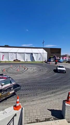A white hatchback drifts in a circular pattern on a paved surface. The car has various decals on its sides, including a panda design. Several orange traffic cones are placed around the car and in a circular pattern behind it, indicating a drift course. In the background, a large white tent structure is visible, with a dark-colored building behind it. The sky is clear and blue.