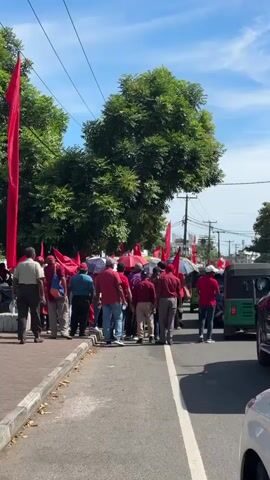 A crowd of people, many dressed in red shirts and carrying red flags, are gathered on a paved road and adjacent sidewalk in Delkanda, Sri Lanka. The scene is outdoors on a bright, sunny day, with a clear blue sky visible above. Lush green trees line the side of the road, and utility poles with overhead wires are present. Some individuals are holding umbrellas, likely for sun protection. Vehicles, including a green tuk-tuk and a dark SUV, are visible on the road beside the crowd. The group appears to be participating in a march, protest, or demonstration, moving in the same general direction away from the camera.