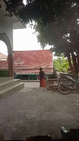 A child in a black shirt and orange skirt stands on a concrete patio, facing away from the camera. The child is holding a white object up to their mouth. To the left of the child are stairs leading up to a white building with an arched entrance. To the right is a motorcycle parked on the patio. In the background, there is a red-tiled roof, green foliage, and a tall tree with dark green leaves. The sky is overcast.