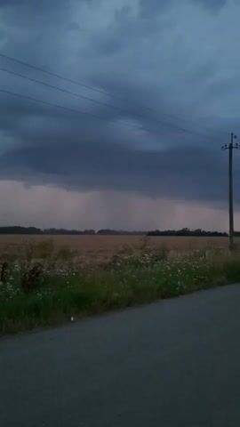 This video captures a rural landscape under a stormy sky. A paved road runs along the bottom of the frame, leading into a field of dry, golden vegetation, likely harvested crops. A line of trees forms a distant horizon. A utility pole with wires stretches across the upper right portion of the image. The sky is dominated by dark, heavy storm clouds, with visible rain shafts falling in the distance, illuminated by a reddish-pink hue, possibly from distant lightning or the setting sun. The lighting suggests dusk or an approaching storm. No people or animals are visible.