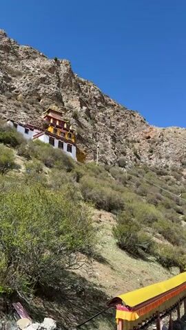 The camera pans across a mountain range in a sunny, daytime scene. The terrain is rocky and grassy, with sparse shrubbery. Visible are several buildings in white, red, and yellow hues, situated on the mountainside. A row of yellow prayer wheels lines a path near the camera. As the camera pans, the vista reveals a vast valley dotted with more buildings, with the mountain range extending into the distance under a clear blue sky with scattered white clouds. The scene suggests a remote, possibly religious or spiritual location, high in the mountains.
