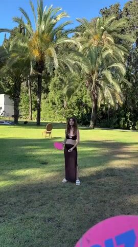 A young woman is playing paddle ball in a grassy backyard with palm trees and other foliage. She is wearing a black bikini top and a sheer black skirt. In the foreground, a large pink paddle with blue lettering is visible, partially obscuring the view of the woman. She is in the process of hitting a ball. The setting appears to be a sunny day.