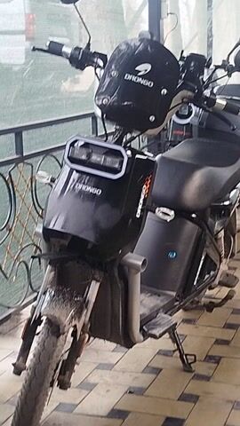 A black electric bicycle or moped is prominently displayed, parked and viewed from its front-left side. The vehicle is predominantly black, featuring a robust frame, black seat, and black handlebars equipped with mirrors on both sides. A black helmet, branded with "DRONGO" in white lettering, hangs from the left handlebar. The brand "DRONGO" is also clearly visible in white and orange lettering on the front light housing, which incorporates a distinctive rectangular LED light unit. A small blue indicator light and a locking mechanism are visible on a side panel of the bike's frame. The front tire and fender show signs of dirt or mud, suggesting recent use.

The background consists of a wall covered with light brown or beige square tiles. In the foreground and midground, a dark metal railing or fence is visible, partially obscuring a blurry view of a structure with a translucent green covering, possibly a greenhouse or a similar outdoor enclosure. The overall lighting is soft and even, indicating either an overcast day or a well-lit shaded outdoor area. The image is oriented vertically, presenting the bike as if it were lying on its side.