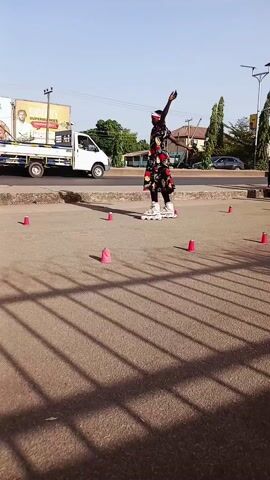 A woman in a long, patterned dress and a red headband is inline skating on a paved area with red cones set in a square pattern. She practices various skating maneuvers as cars pass on the adjacent road. Another person, wearing a white t-shirt and black pants, is also inline skating nearby. The scene takes place outdoors on a sunny day, with buildings, trees, and street signs visible in the background. The shadow of a fence runs diagonally across the bottom of the frame.