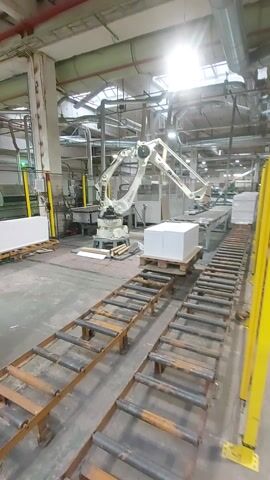 The video shows a robotic arm operating in a factory setting. The robotic arm, painted white and red, is positioned above a pallet of what appear to be stacked white boxes or paper reams. It is actively moving and manipulating the boxes, suggesting an automated packaging or sorting process. In the background, industrial equipment, including large pipes and ventilation systems, are visible. A conveyor belt system, made of metal rollers, runs alongside the robotic arm. Yellow safety barriers are also present, indicating a controlled work environment. The lighting suggests overhead factory lights, with a bright glare visible in the upper left portion of the frame. The overall scene depicts an automated industrial operation.