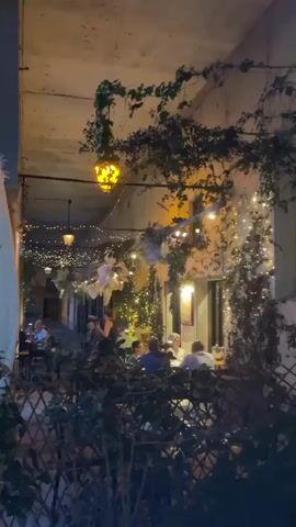 The video opens with a close-up of an outdoor dining area at dusk. String lights and lanterns illuminate the space, casting a warm glow on ivy-covered walls and tables where people are seated, eating and conversing. The camera then pans left, revealing a large, weathered building with arched walkways on the ground floor, leading to a courtyard filled with parked cars. A tall, old tower, also part of the building, stands prominently against the twilight sky. The scene suggests a relaxed evening atmosphere in a historic setting, possibly a town square or a restaurant courtyard.