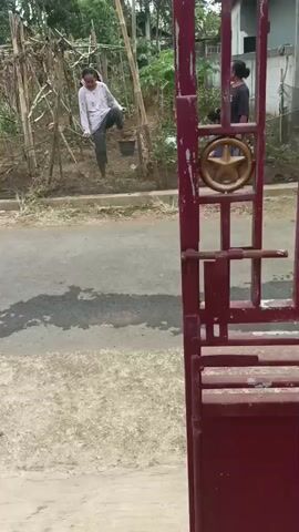 A girl is swinging on a makeshift swing. A woman is standing and watching her. A person walks by in the background. The setting appears to be a rural or suburban area with vegetation and some simple structures.