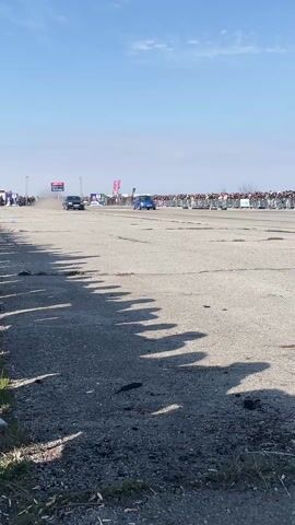 The video shows two cars drag racing on an asphalt course, with a crowd of people lining the side of the track. The cars, a dark colored Volkswagen Golf and a blue Volkswagen Beetle, accelerate quickly side-by-side. The Beetle edges ahead, and the cars race down the course, kicking up dust. The camera then tracks the Beetle as it continues down the race course on its own. The sky is bright blue with some clouds. Some tree shadows stretch onto the pavement.