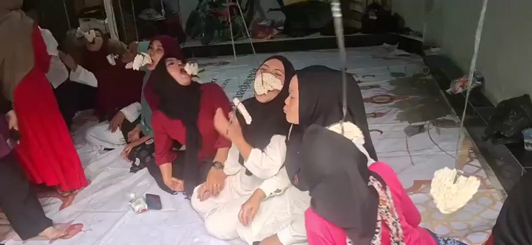 A group of young women and girls are sitting on a white sheet on the floor, facing forward. Each of them has a cracker attached to a string hanging from above, and they are attempting to eat the cracker without using their hands. Some of the participants are laughing and appear to be enjoying the challenge. The environment appears to be indoors, with a tiled floor and some furniture visible in the background. The overall atmosphere suggests a fun and playful gathering, possibly a party or a cultural event.