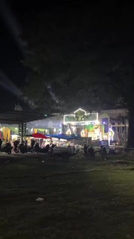 A night scene is depicted. In the foreground, a grassy area is visible, with some discarded items on the ground. In the middle ground, several motorcycles are parked. Behind the motorcycles, there are market stalls with awnings and tables, where people are seated. Further back, a stage is set up with bright lights and decorations, including a large, illuminated sign with a dome shape. Some individuals are standing on the stage. The overall atmosphere appears to be that of an outdoor gathering or event at night.