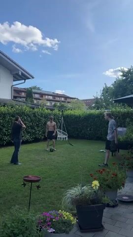 Three young men are playing soccer in a backyard. The man on the left is wearing a dark shirt and jeans. The man in the middle is shirtless and wearing black shorts. The man on the right is wearing a gray and white checkered shirt and shorts. The man on the right kicks the soccer ball into the air. The man on the left attempts to kick the ball but misses. The man in the middle watches the ball. The yard is green and surrounded by a tall hedge. A house is visible in the background. There are flowers in pots in the foreground.