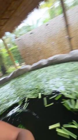 A woman is shown bathing in a pond or tank, seemingly at a spa or resort. The water is filled with cut, green plants floating on the surface. The woman is wearing a swimsuit and has her hair in a bun. She looks at the camera, smiles, and turns her head, enjoying her bath. A thatched roof covers the area, and greenery surrounds the pool. There is a sign with text behind her.