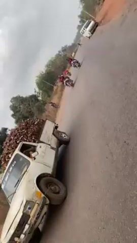 The video shows a road in a rural area. In the first shot, a white pickup truck full of what appears to be coconuts is parked on the side of the road. Two motorcyclists pass by. In the second shot, the camera pans to a dirt area beside the road. The third and fourth shots are from the perspective of someone riding on a motorcycle, following another motorcycle ahead. The road is paved and lined with trees. The sky is overcast.
