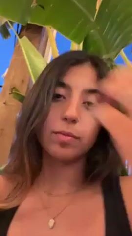 Here is a detailed description of the video:

The video features a young woman with medium-length brown hair, a fair complexion, and brown eyes. She is wearing a black tank top and a delicate gold necklace with a pendant. She also has multiple rings on her fingers, including a band on her ring finger. The woman is holding a gold hair clip in her hand and then puts it between her teeth.

The setting appears to be outdoors, possibly on a patio or in a garden. Large green leaves are visible in the background, suggesting the presence of tropical plants. The lighting suggests it could be evening, with a hint of blue sky visible through the foliage.

The woman is the main focus of the video, and she seems to be talking directly to the camera, although the content of her speech is unclear. Her expression is somewhat neutral.