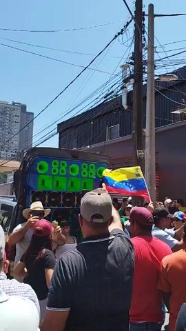 A group of people are gathered in a street, waving Venezuelan flags. They are standing near a building with a sign that reads "COMERCIALIZADORA MR QUESOS". A large speaker system is also present, playing music. The people appear to be celebrating or protesting. The sky is blue and the weather is sunny. A lot of power lines overhead the crowd.
