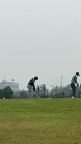 Two men are on a golf course. The man on the left is wearing a striped shirt and a hat. The man on the right is wearing a green shirt and a hat. Both men are holding golf clubs. The man on the left is preparing to swing. The man on the right is looking down. There are trees and buildings in the background. The sky is overcast.