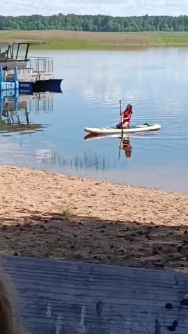 A person is paddleboarding on a calm lake on a sunny day. The person is wearing an orange life vest and is using a paddle to move across the water. In the background, there is a dock with a boat and a forested shoreline. The foreground shows a sandy beach and some wooden structures. The water is clear and reflects the sky and surrounding scenery. There are some faint reeds visible in the water near the shore. The weather appears to be warm and pleasant, suitable for outdoor water activities. The overall scene depicts a peaceful and recreational setting in Belarus.