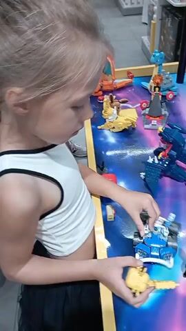 A young girl with blonde hair is playing with toys on a table. She is wearing a white tank top and black pants. She is holding a yellow toy in her hands and appears to be interacting with other toys on the table, which include various colorful figures and structures. The table has a galaxy-themed surface. Another child can be seen in the background, also playing with toys. The environment appears to be indoors, possibly a store or a play area.