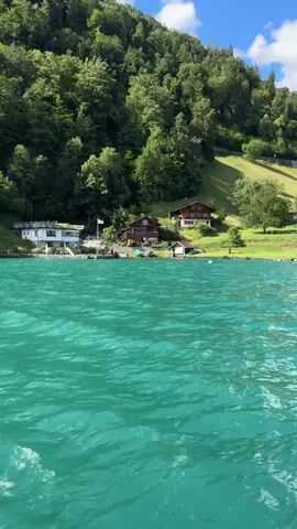 The video captures a scenic view from a body of water, likely a lake, with turquoise-colored water in the foreground. The camera appears to be moving, suggesting it is on a boat. In the middle ground, a shoreline is visible, lined with several buildings, some appearing to be houses with red roofs, and others more modern structures. A white boat is docked near one of the buildings. Behind the buildings, a lush green hillside rises steeply, covered in dense trees. The sky above is blue with scattered white clouds. The overall impression is one of a serene natural landscape with human habitation nestled within it.