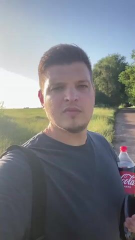 A man is holding a bottle of Coca-Cola and walking down a path in a rural area. The sun is shining and the sky is blue. There are trees and grass on either side of the path. The man is wearing a dark t-shirt and a backpack. He is looking at the camera and appears to be talking.